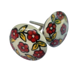 May include: Two ceramic cabinet knobs with a cream-colored base. Each knob is decorated with red flowers, green leaves, and yellow accents. The knobs have a silver-colored screw for installation. These decorative knobs are perfect for adding a touch of floral charm to furniture.