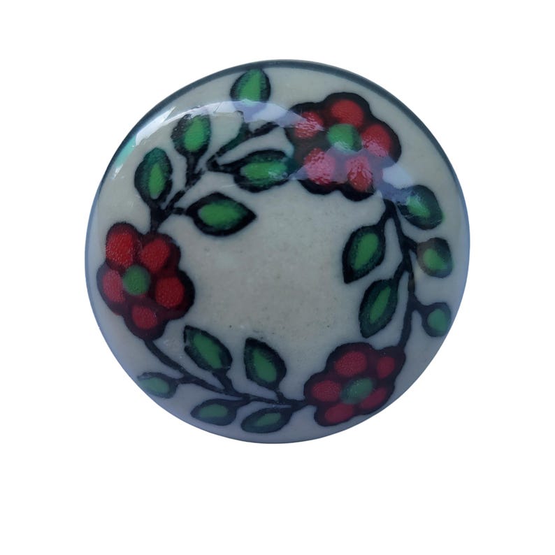 May include: A circular ceramic cabinet knob in off-white, decorated with a floral pattern. The design includes red flowers with green centres and green leaves on black stems. Suitable for furniture or decorative use.