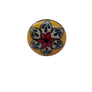 Op de afbeelding: Ronde, gele keramische kastknop met een bloemmotief. De bloem heeft een rood hart, witte bloemblaadjes en donkergroene bladaccenten. De knop is een decoratief hardware-element.
