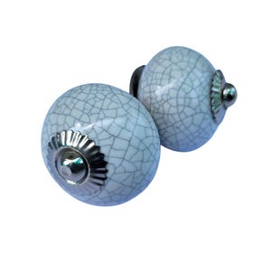 May include: Two white ceramic cabinet knobs with a crackle glaze pattern. Each knob has a silver-colored metal base and a small, round silver accent. The knobs are spherical and designed for furniture.