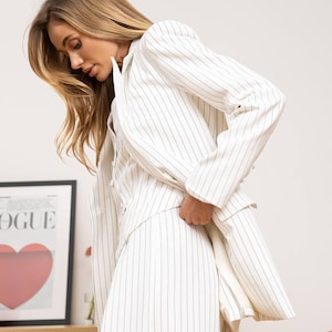 May include: White pinstripe blazer and matching skirt. The outfit features thin black vertical stripes on a white background. The blazer has a classic cut with long sleeves. The skirt is a midi length. The outfit is part of a fashion collection.