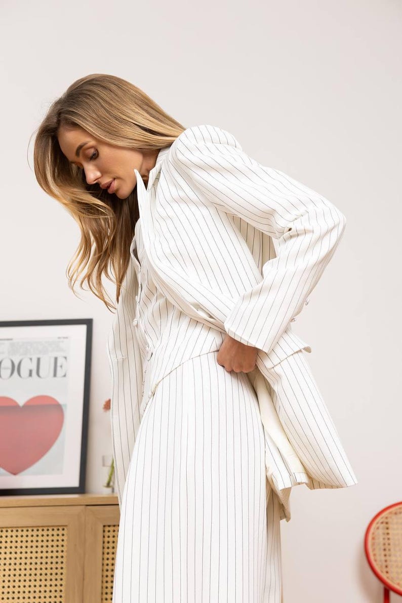 May include: White pinstripe blazer and matching skirt. The outfit features thin black vertical stripes on a white background. The blazer has a classic cut with long sleeves. The skirt is a midi length. The outfit is part of a fashion collection.