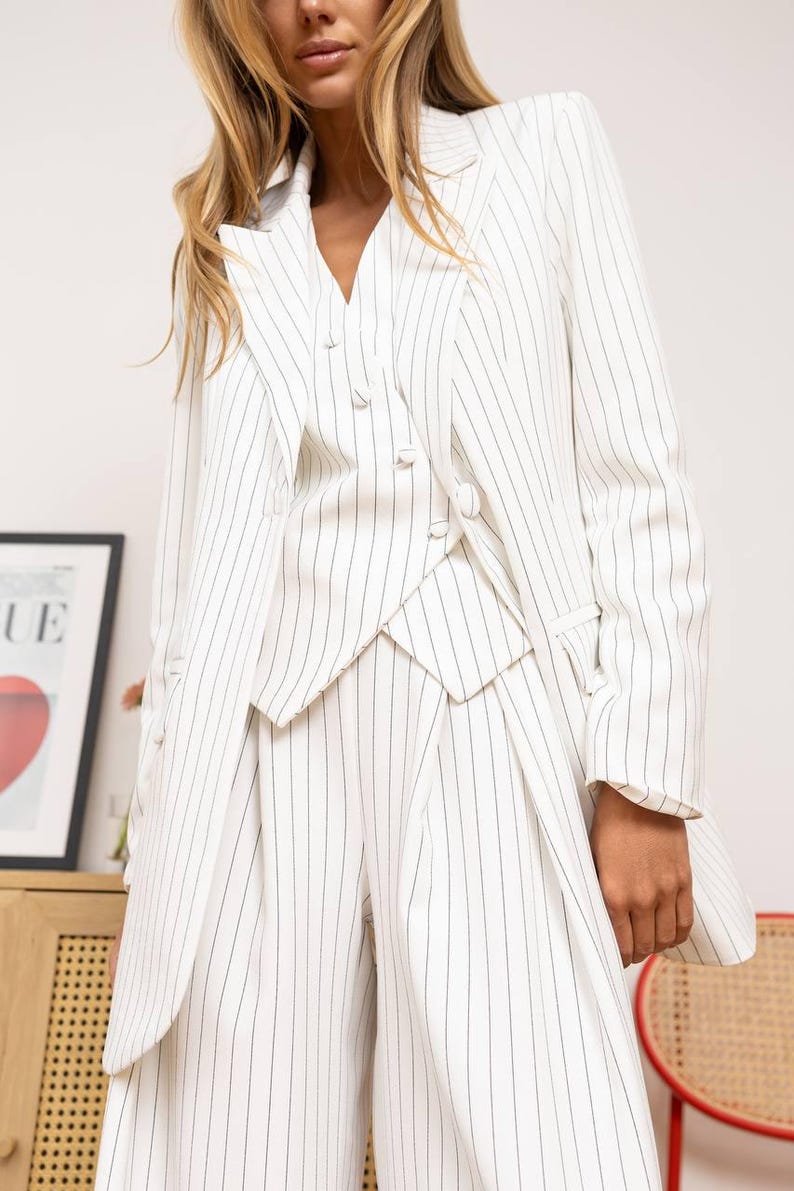 May include: White pinstripe blazer, vest, and wide-leg pants. The suit features a double-breasted blazer with notched lapels, a matching vest, and high-waisted pants. The pinstripes are a dark gray color.