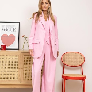 May include: A three-piece pink suit, including a blazer, vest, and wide-leg pants. The suit is worn by a person standing in front of a wooden cabinet, a red chair, and a framed "Vogue" poster with a heart design.
