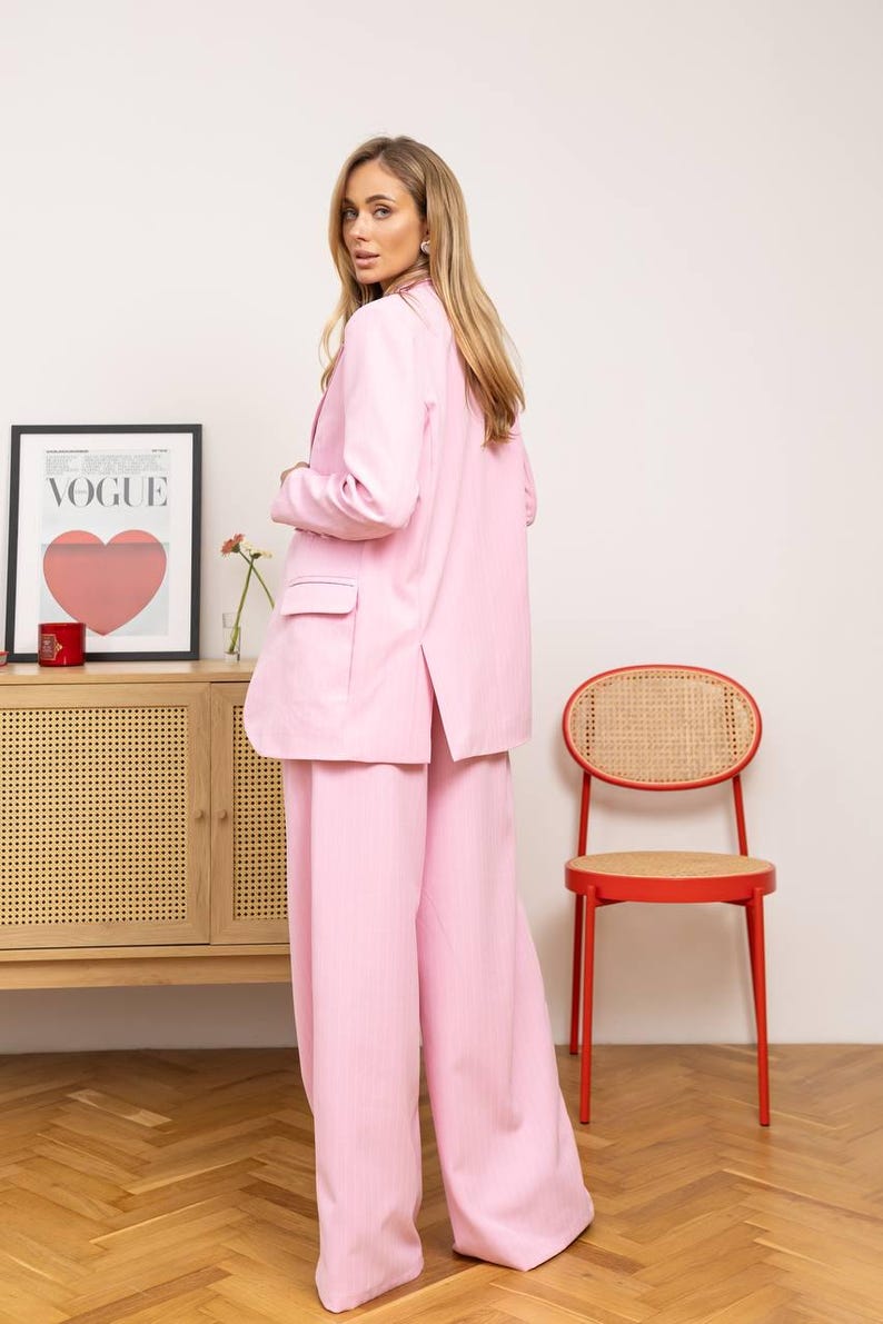 May include: A light pink suit, including a blazer and wide-leg pants. The blazer has pockets and a split back. A red chair with a woven back and a framed "VOGUE" poster with a heart graphic are in the background.