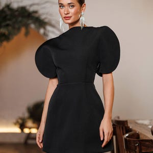 May include: A black mini dress with dramatic, oversized puff sleeves. The dress features a high neckline and a fitted waist, creating a structured silhouette. The model is wearing silver dangling earrings. The background is a neutral color.