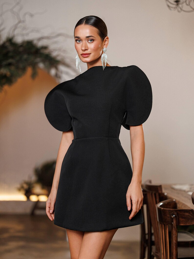 May include: A black mini dress with dramatic, oversized puff sleeves. The dress features a high neckline and a fitted waist, creating a structured silhouette. The model is wearing silver dangling earrings. The background is a neutral color.