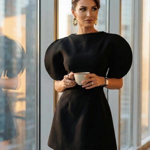 May include: A black dress with short, puffed sleeves and a flared skirt. The woman is holding a white teacup with a gold rim. She is wearing gold earrings and a gold watch. The dress is a classic, elegant style.