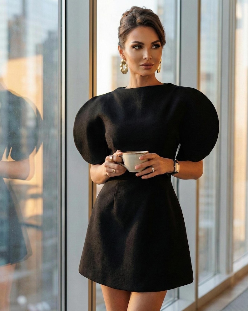 May include: A black dress with short, puffed sleeves and a flared skirt. The woman is holding a white teacup with a gold rim. She is wearing gold earrings and a gold watch. The dress is a classic, elegant style.