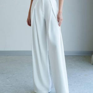 May include: White, high-waisted wide-leg trousers. The trousers feature a tailored design with front pleats and a button closure. The fabric appears smooth and opaque. The trousers are worn with a light pink top and grey pointed-toe shoes.