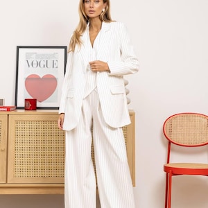 May include: A white pinstripe suit, including a blazer and wide-leg pants. The blazer has pockets and a notched collar. The outfit is worn by a person. A framed "VOGUE" poster with a red heart is visible in the background.