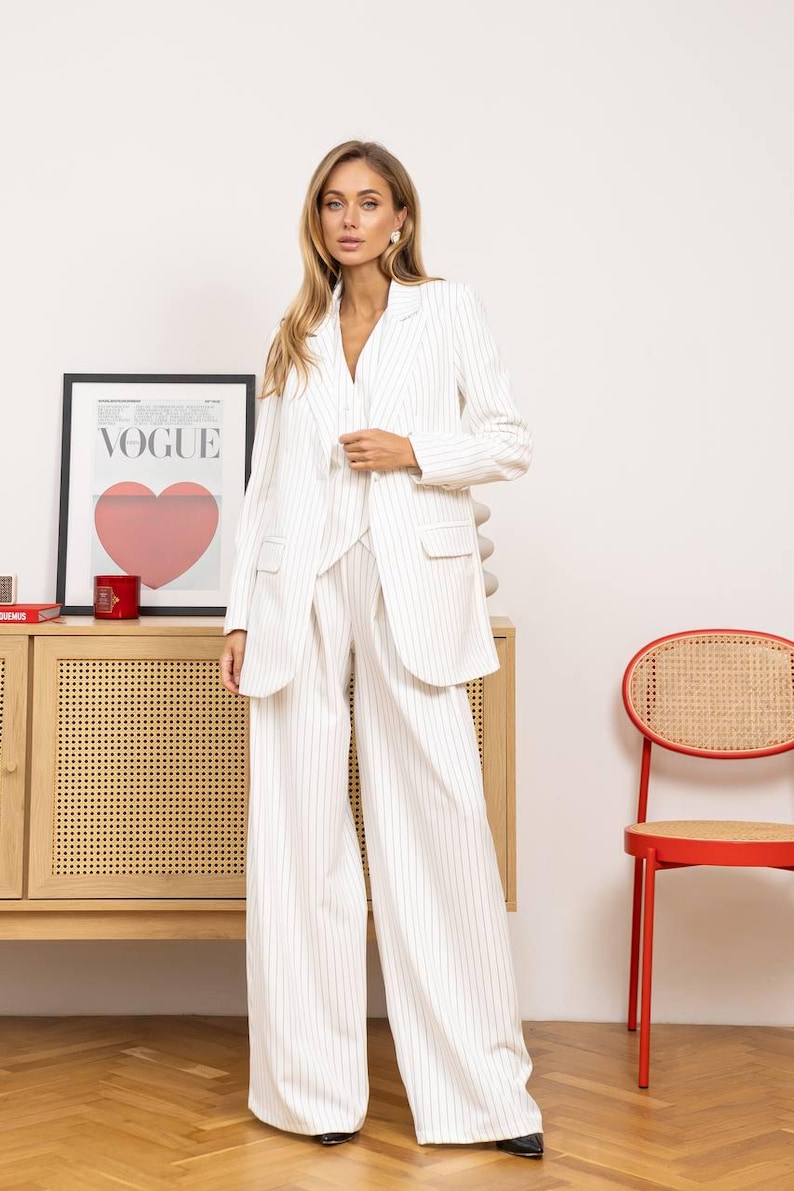 May include: A white pinstripe suit, including a blazer and wide-leg pants. The blazer has pockets and a notched collar. The outfit is worn by a person. A framed "VOGUE" poster with a red heart is visible in the background.