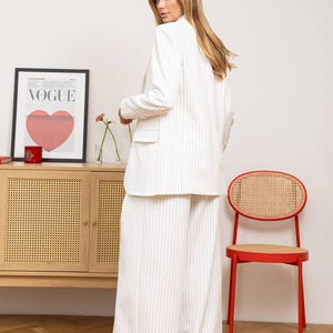 May include: White pinstripe blazer and wide-leg pants. The outfit is part of a matching set. A red chair and a wooden cabinet with a Vogue poster are in the background.