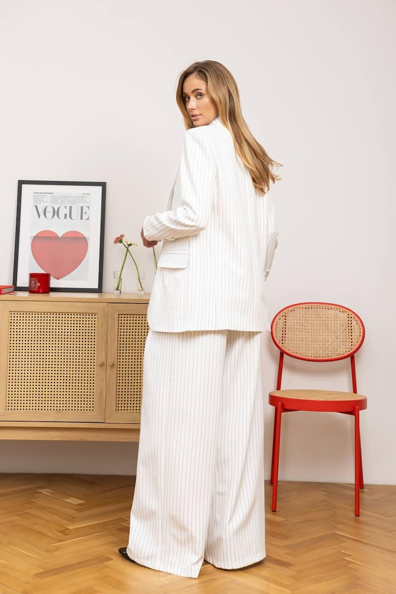 May include: White pinstripe blazer and wide-leg pants. The outfit is part of a matching set. A red chair and a wooden cabinet with a Vogue poster are in the background.