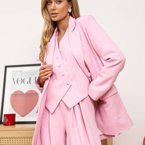 May include: A light pink three-piece suit, including a blazer, vest, and wide-leg pants. The blazer has a notched lapel and the vest has buttons. The suit is made of a textured fabric. A framed Vogue poster is in the background.