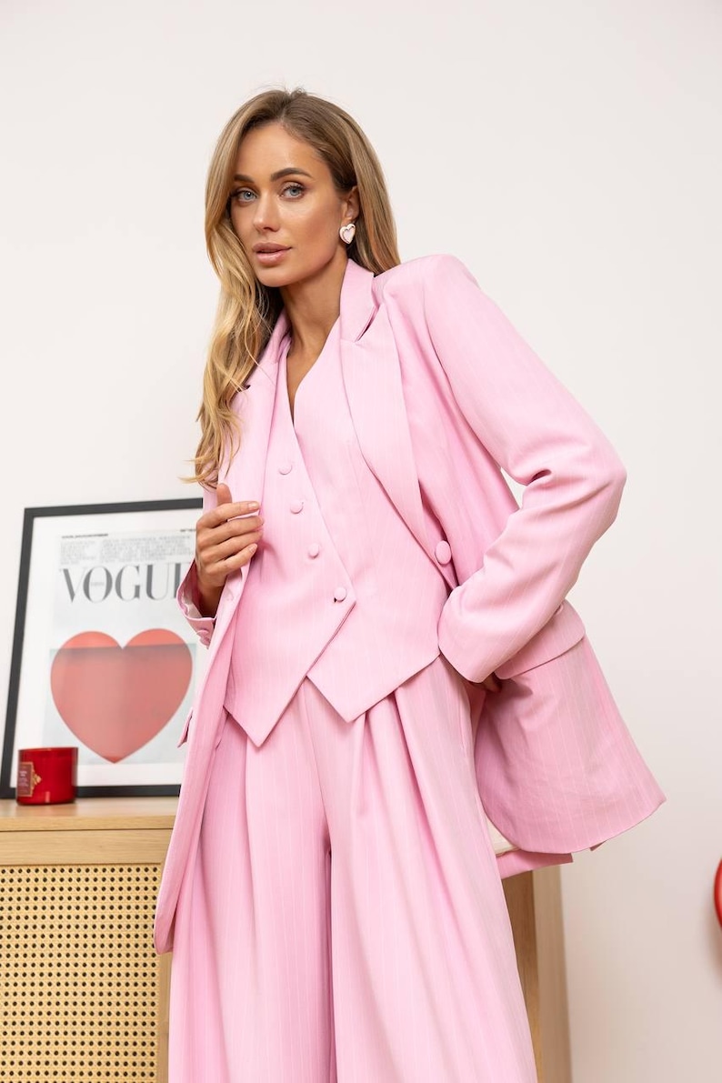 May include: A light pink three-piece suit, including a blazer, vest, and wide-leg pants. The blazer has a notched lapel and the vest has buttons. The suit is made of a textured fabric. A framed Vogue poster is in the background.