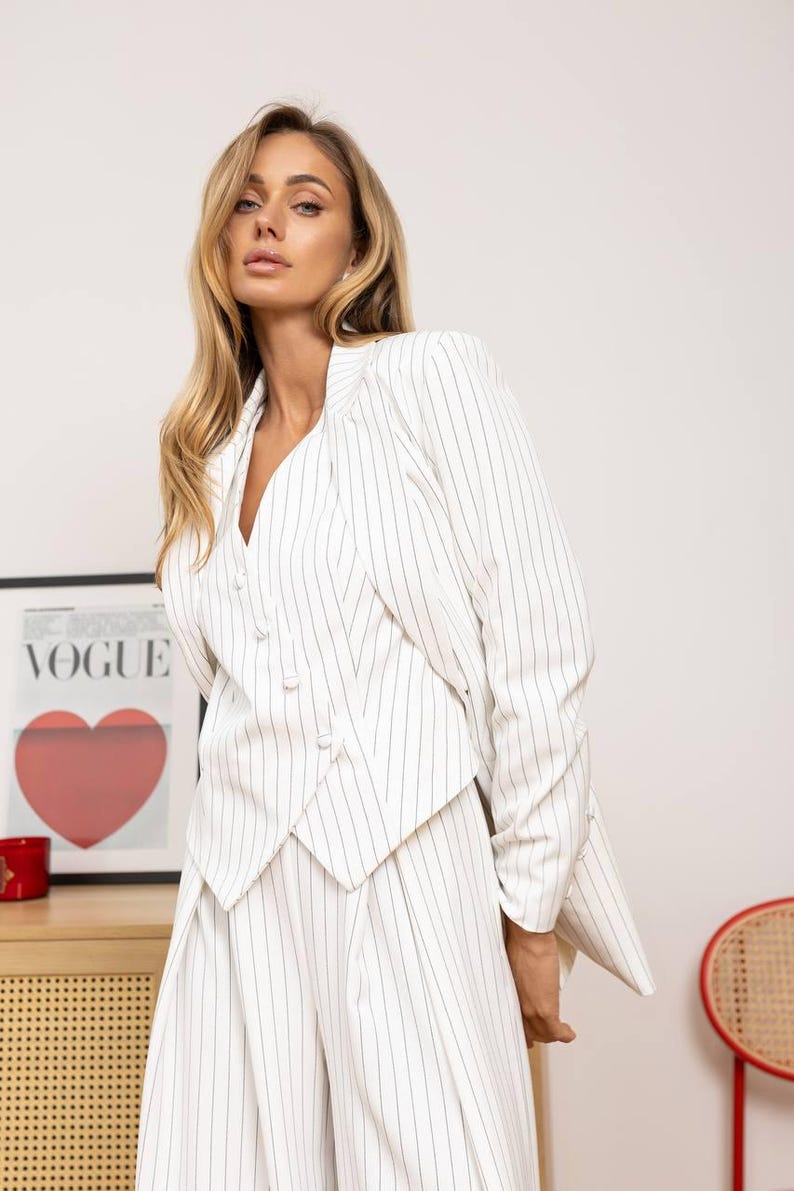 May include: White and black pinstripe blazer and matching pants. The blazer has a unique button design and a V-neck. The outfit is part of a fashion collection. A Vogue poster with a red heart is in the background.