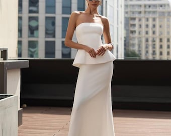 May include: A strapless white dress with a peplum top and a long, fitted skirt. The dress is worn by a person wearing sunglasses. The background features a cityscape with modern buildings and a wooden deck.