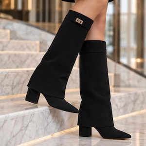 Könnte beinhalten: Ein Paar schwarze kniehohe Stiefel mit einem breiten, umgeschlagenen Schaftabschluss. Die Stiefel verfügen über eine spitz zulaufende Zehenpartie, einen Blockabsatz und ein dekoratives, goldfarbenes rechteckiges Schnallendetail am Schaft.