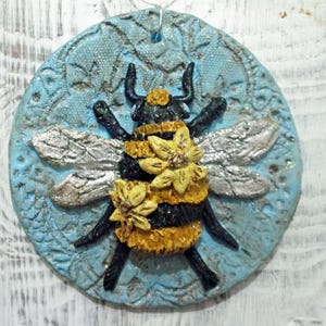 May include: A round, blue and silver ornament with a 3D bumble bee design. The bee has black and yellow stripes, silver wings, and yellow flowers.