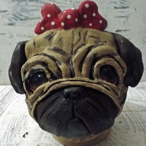 May include: A ceramic pug dog figurine with a red and white polka dot bow on its head. The figurine has a realistic appearance with brown eyes and a wrinkled face.