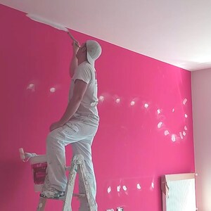 Può includere: Una persona sta dipingendo una parete rosa con vernice bianca usando un rullo e un pennello. La persona è in piedi su una scala a pioli. La parete è parzialmente dipinta e presenta macchie bianche di vernice.