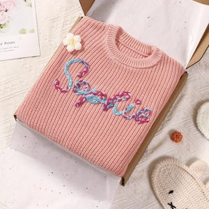 Può includere: Un maglione rosa lavorato a maglia con il nome "Sophie" ricamato con filo blu e rosa. Sono visibili anche un fiore bianco e una piccola zucca arancione. Il maglione è piegato all'interno di una scatola di cartone.
