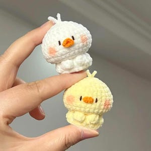 May include: Two crocheted duck figures. One is white, the other is yellow. Both have orange beaks, pink cheeks, and small black eyes. The ducks are handmade and have a fluffy texture. They are held between fingers.