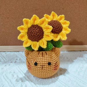 May include: Crocheted sunflower arrangement in a cat-shaped pot. The sunflowers have yellow petals and brown centers. The pot is orange with black eyes and pink cheeks. The arrangement is on a white, textured surface.