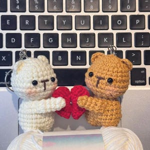 May include: Two crocheted bear keychains, one cream and one tan, holding a red heart. The bears have black eyes and brown noses. The keychains are on a laptop keyboard, with a ball of white yarn in the foreground.