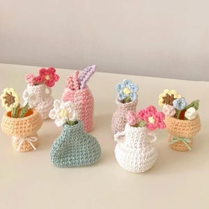 Puede incluir: Una colección de jarrones de ganchillo hechos a mano en varios colores, cada uno con una flor de ganchillo. Los jarrones son en tonos de blanco, rosa, azul y amarillo. Algunos jarrones tienen asas y algunos están atados con una cinta. Las flores son rosas, amarillas y azules.