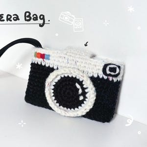May include: A black and white crocheted camera bag with a black wrist strap. The bag features a circular lens detail, a small square viewfinder, and a rainbow-colored accent on the top edge. The text "Camera Bag." is printed above the item.