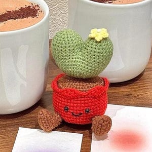 Puede incluir: Dos tazas blancas llenas de una bebida marrón, cada una cubierta con una línea marrón oscuro. Un cactus de ganchillo con una flor amarilla se encuentra frente a las tazas. Una maceta roja de ganchillo con una cara sonriente sostiene el cactus. También hay dos pequeñas tarjetas con degradados morados y rojos.