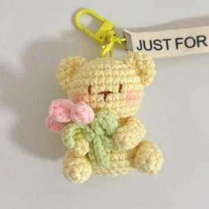 Puede incluir: Un llavero de oso de peluche amarillo tejido a ganchillo que sostiene un ramo de flores rosas y verdes. El llavero tiene un cierre amarillo y una etiqueta blanca que dice "JUST FOR YOU". El oso está hecho de hilo suave.