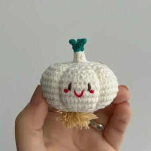 May include: A handmade, crocheted garlic bulb with a smiling face. The white bulb has a green stem and a yellow fringe at the base. The face features red cheeks and a red smiling mouth. This whimsical item is a unique decorative piece.