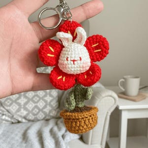 May include: A handmade crochet keychain featuring a red flower with a white bunny face at the center. The bunny has pink cheeks and a small red mouth. Below the flower is a green cactus in a brown pot. The keychain has a silver clasp.