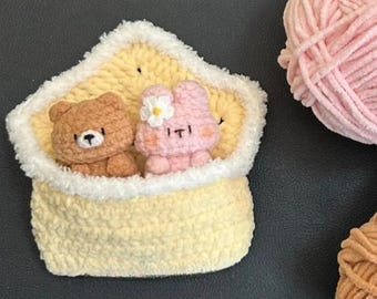 Puede incluir: Una pieza de ganchillo hecha a mano con un oso marrón y un conejito rosa con una flor, metidos en una bolsa amarilla con ribete blanco. El fondo incluye ovillos de lana rosa, amarilla y marrón.