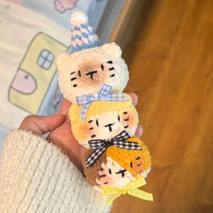 May include: A vertical stack of three crocheted plush animal heads. The top head is cream-colored with a blue and white party hat. The middle head is yellow, and the bottom head is brown and orange. Each head features a small decorative fabric bow.