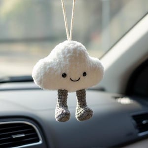 Könnte beinhalten: Ein flauschiger, weißer Wolken-Autoanhänger mit einem lächelnden Gesicht und grauen gehäkelten Beinen. Die Plüschdekoration hängt an einer Schnur und verleiht dem Innenraum des Fahrzeugs eine skurrile Note. Ein niedliches und fröhliches Accessoire.