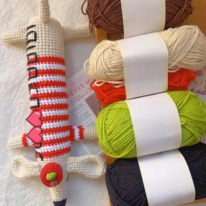 May include: A handmade crochet giraffe toy with red and white striped details, a red apple, and a black and white face. Beside the giraffe are several skeins of yarn in brown, cream, red, green, and black.