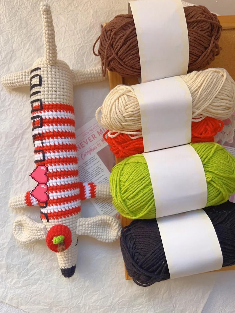 May include: A handmade crochet giraffe toy with red and white striped details, a red apple, and a black and white face. Beside the giraffe are several skeins of yarn in brown, cream, red, green, and black.