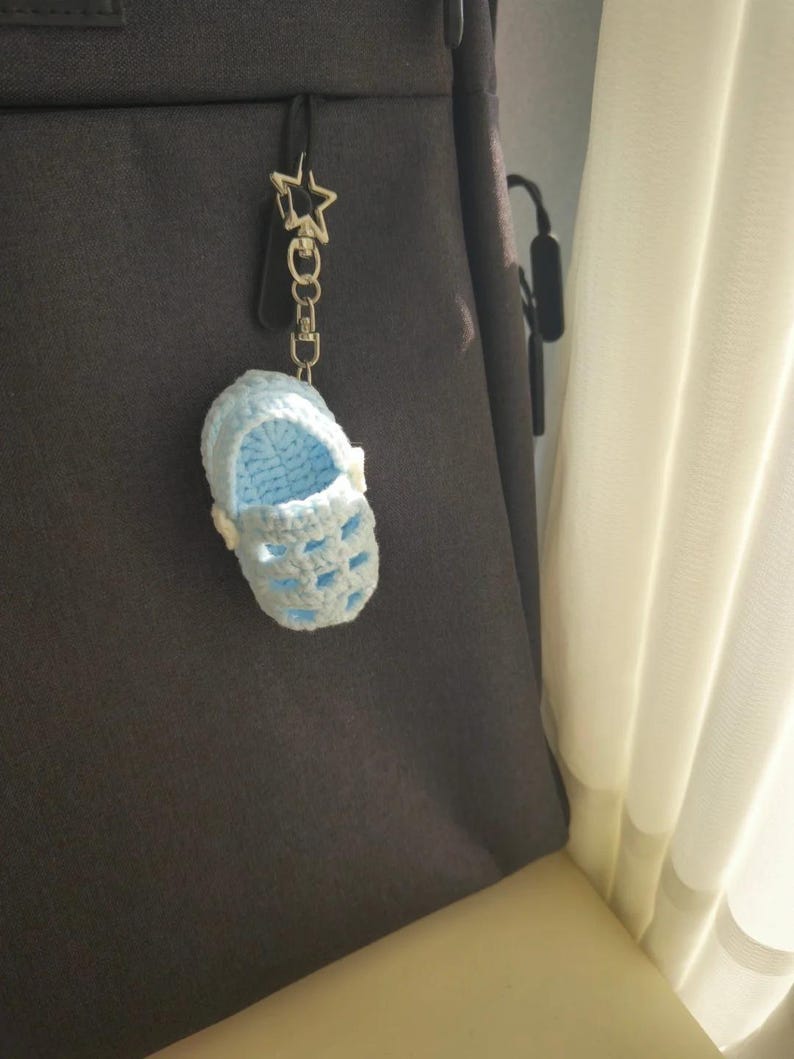 Op de afbeelding: Een lichtblauwe gehaakte babyschoen-sleutelhanger met een stervormige metalen sluiting. Het miniatuurschoentje heeft een uitgesneden ontwerp, een ronde neus en een klein wit knoopdetail aan de zijkant, hangend aan een zilverkleurige metalen ketting.