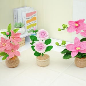 May include: Three crochet flower arrangements in small pots. The flowers are pink and white, with green leaves. The pots are a light brown color. The flowers are arranged on a white surface.