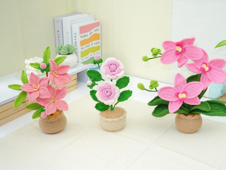 May include: Three crochet flower arrangements in small pots. The flowers are pink and white, with green leaves. The pots are a light brown color. The flowers are arranged on a white surface.