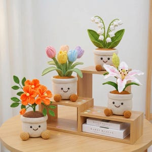 May include: Four crocheted potted plants with smiling faces on the pots. One pot holds orange flowers, one holds pastel tulips, one holds white lily of the valley, and one holds a pink and white lily. The pots have small round feet and brown tops.