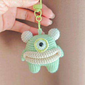 May include: A green crocheted monster keychain with a single large eye and a white mouth. The keychain is attached to a green and white plastic key ring.