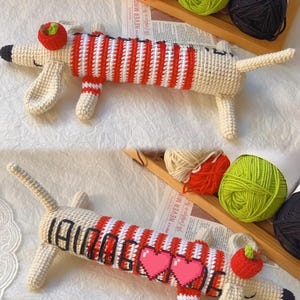 May include: Crocheted dachshund dog toy in cream, red, and white. The dog has a red and white striped body, a red apple on its head, and black accents. The word "DACHSHUND" is written on the dog's body with two pink pixelated hearts.