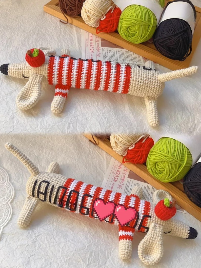 May include: Crocheted dachshund dog toy in cream, red, and white. The dog has a red and white striped body, a red apple on its head, and black accents. The word "DACHSHUND" is written on the dog's body with two pink pixelated hearts.