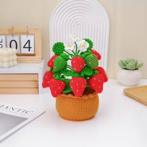 Pode incluir: Uma planta de morango de crochê feita à mão em uma cesta tecida. A planta apresenta morangos vermelhos brilhantes com detalhes de sementes brancas, folhas verdes e flores brancas. A cesta é de cor marrom quente, em um fundo neutro.