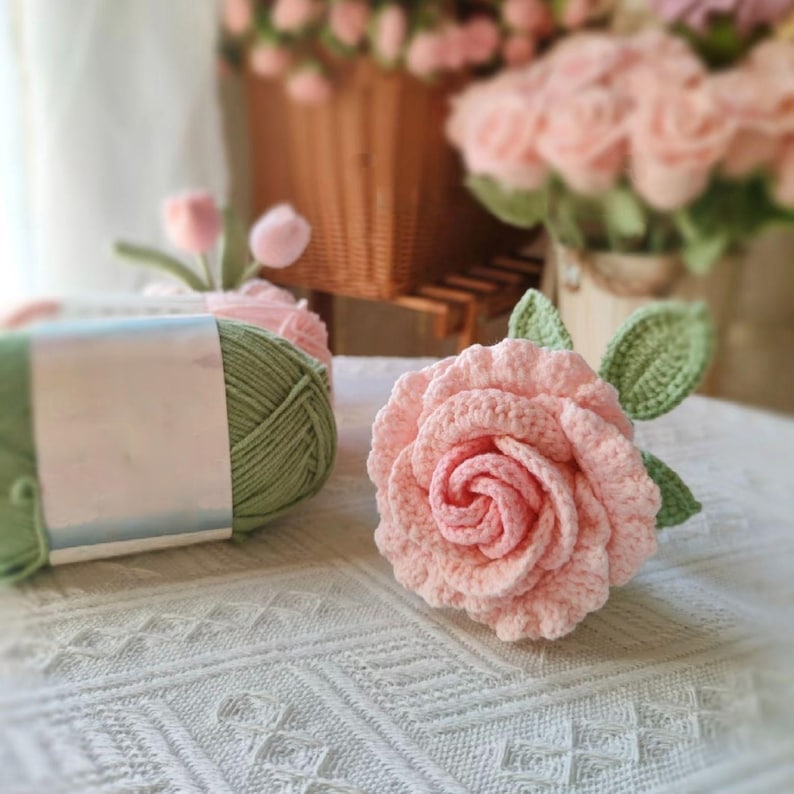 May include: A crocheted pink rose with green leaves. The rose is sitting on a white crocheted tablecloth. There is a ball of green yarn and a ball of pink yarn in the background.